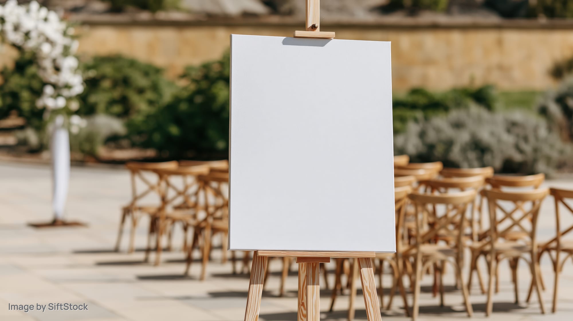 Blank white canvas on wooden easel in outdoor wedding setup with chairs and floral decor