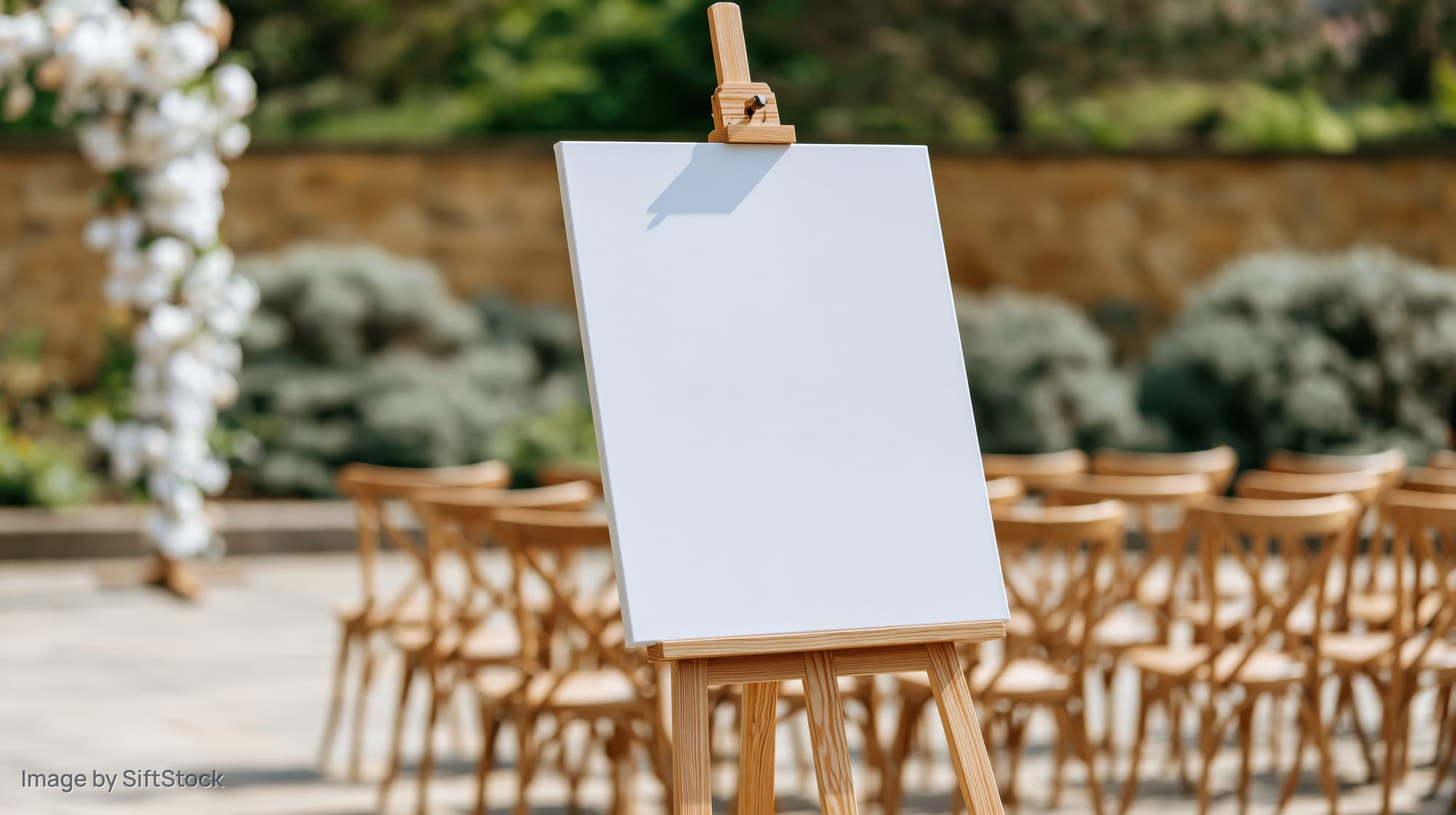 Blank white canvas on wooden easel in outdoor wedding ceremony setup with chairs and floral arch
