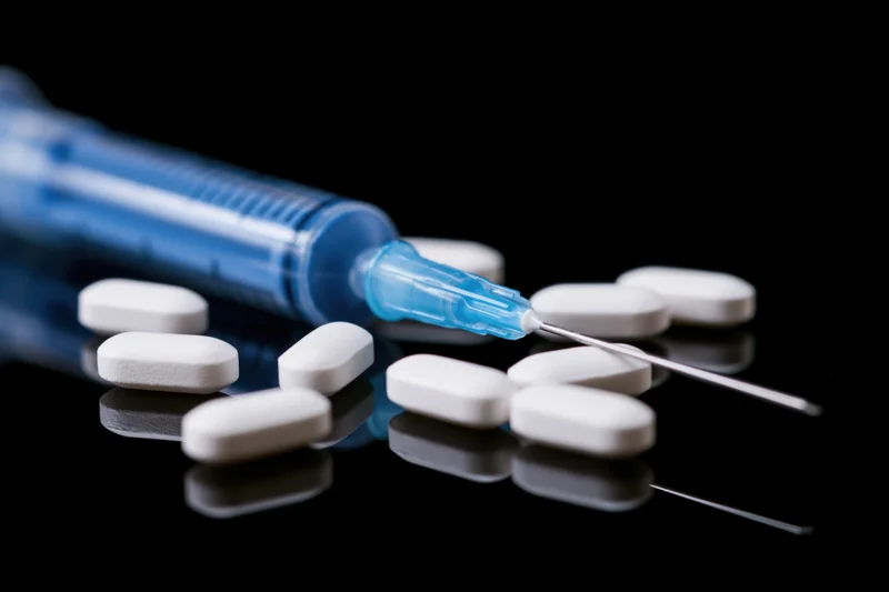 Blue medical syringe with needle lying beside white pills on a reflective black surface