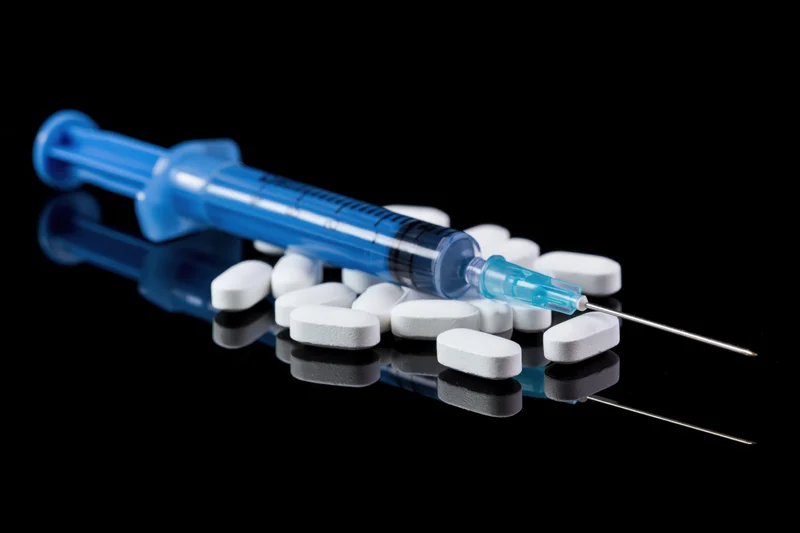 Blue medical syringe with needle lying on glossy black surface among white tablets