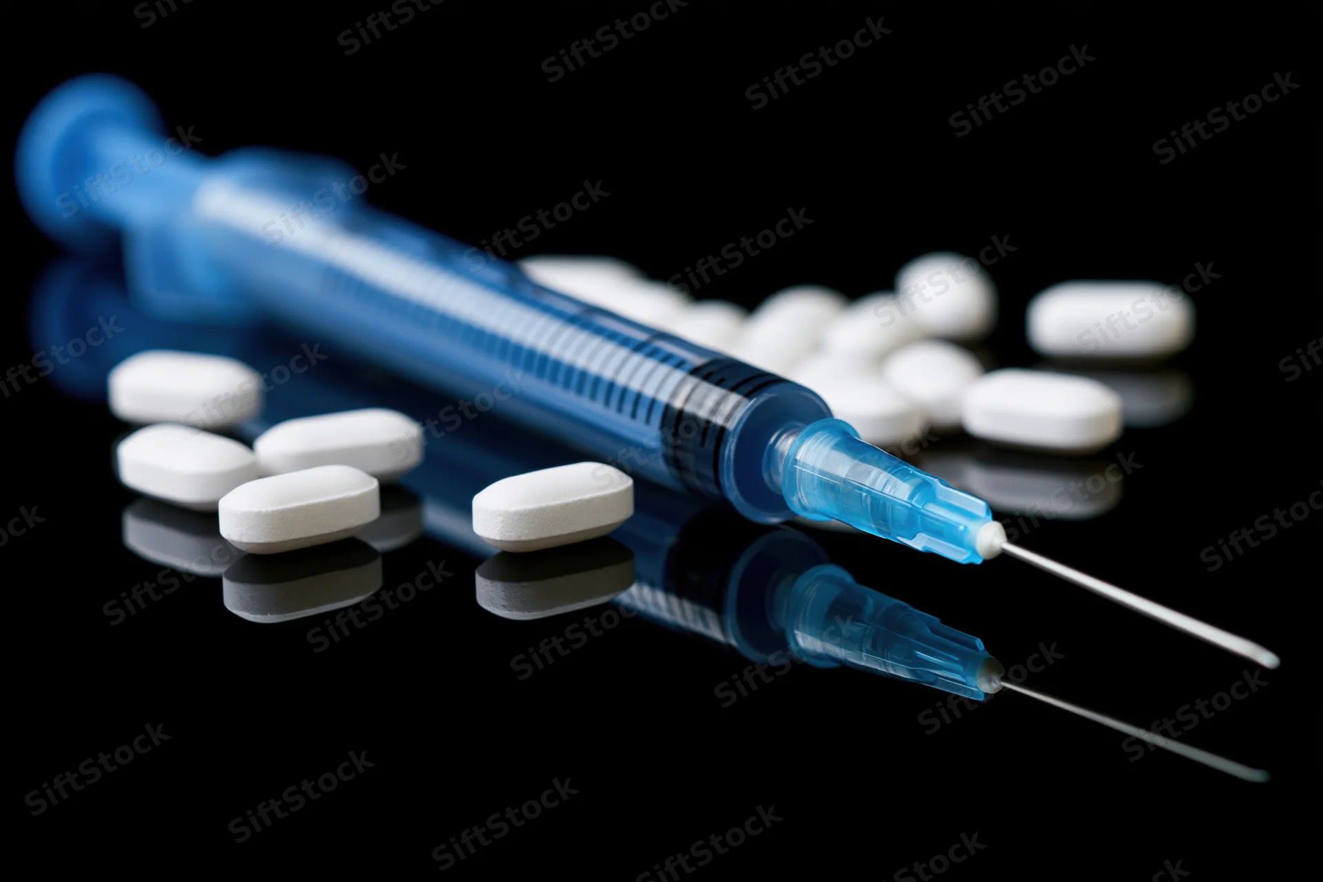 Blue medical syringe with needle and white pills on reflective black surface