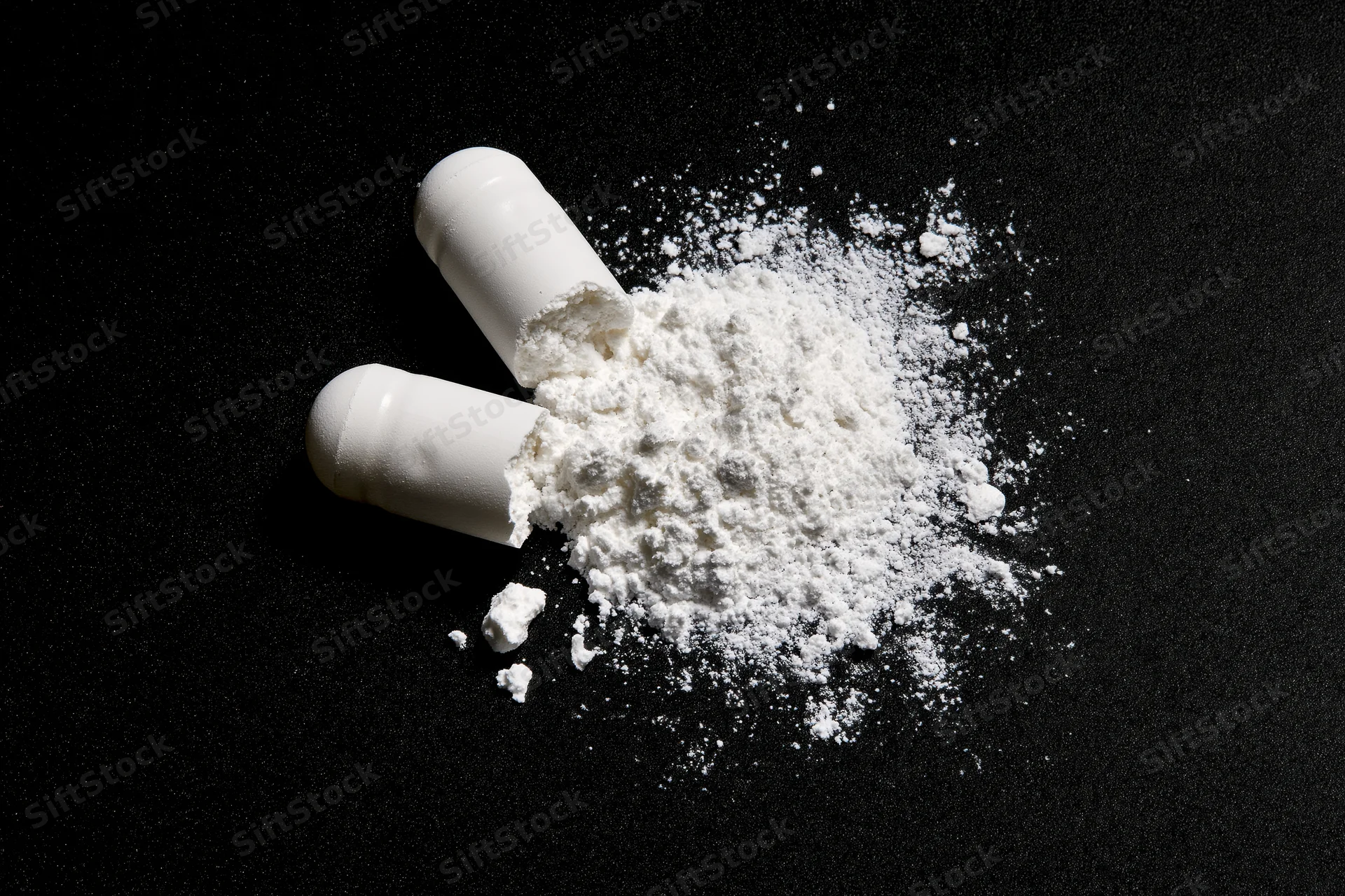 Broken white pharmaceutical capsule split open spilling powder on black surface