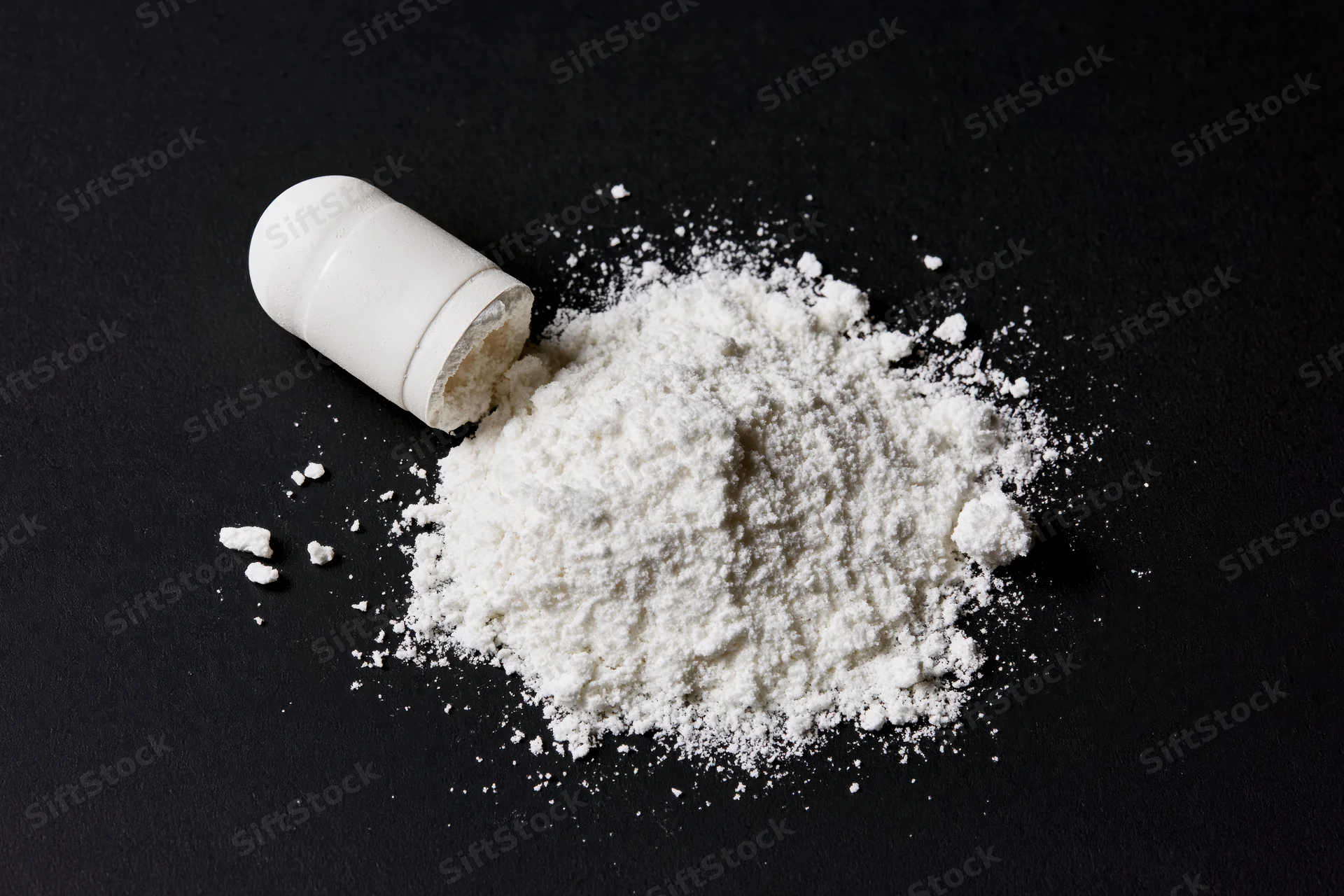 Broken white pharmaceutical capsule spilled onto black surface, revealing fine white powder inside