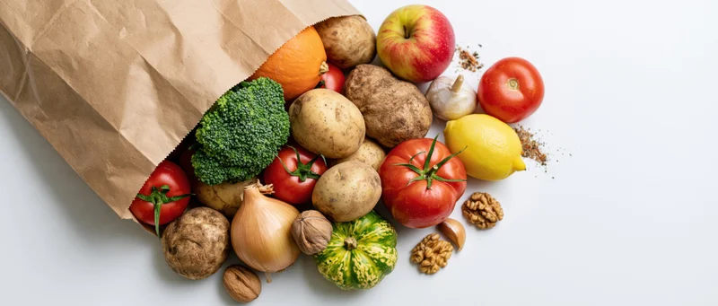 Brown paper grocery bag tipped over spilling fresh vegetables and fruits onto white surface