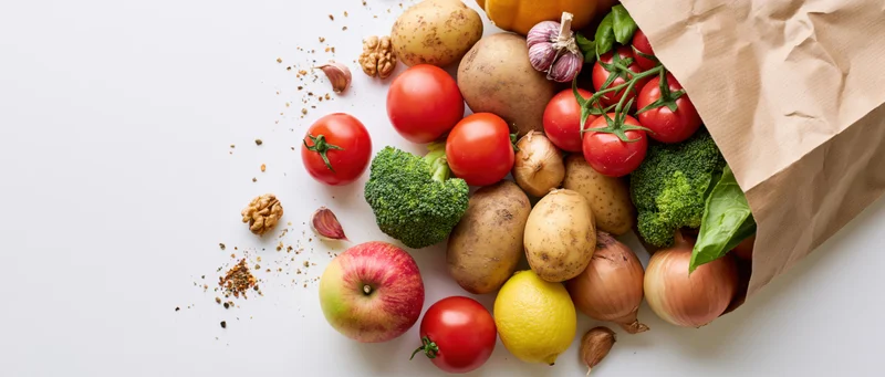 Brown paper grocery bag tipped over spilling fresh vegetables and fruits onto white surface