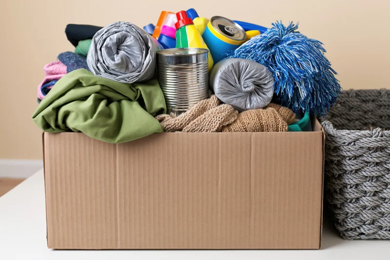 Cardboard donation box overflowing with mixed household items including clothing, canned food, and cleaning supplies on a table