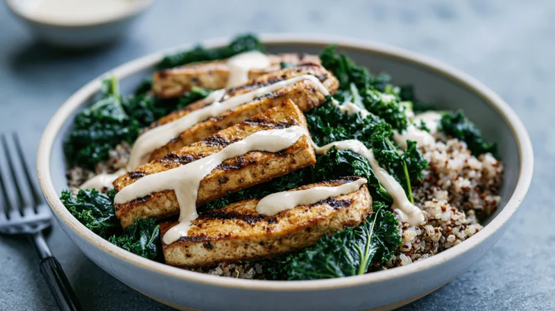 Grilled tofu slabs with sautéed kale and quinoa in a ceramic bowl, drizzled with sauce, on a gray surface with a fork beside it