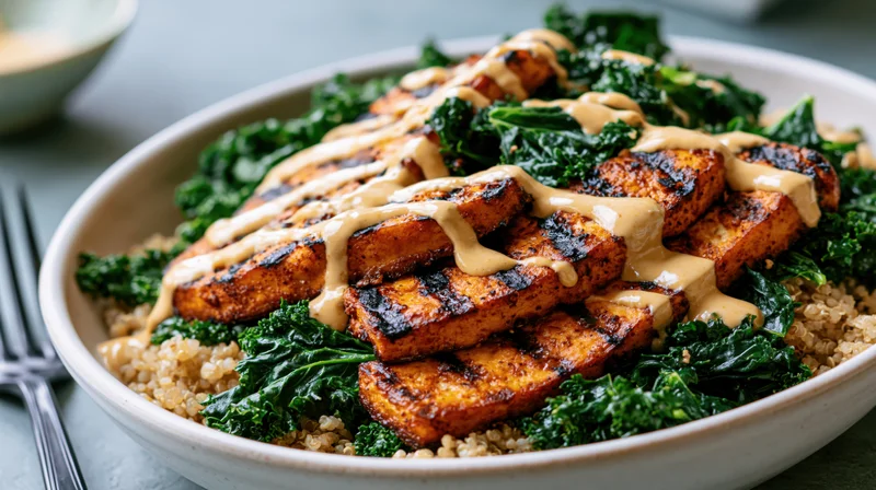 Grilled tofu slabs with sautéed kale and quinoa in a ceramic bowl, drizzled with sauce, on a table with a fork beside it