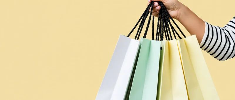 Hand holding multiple paper shopping bags with black rope handles against a yellow background