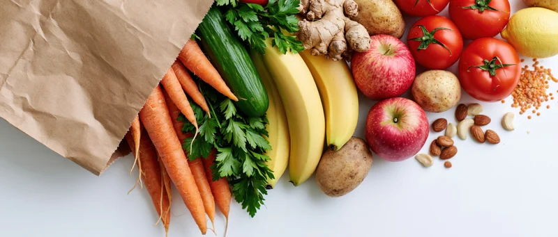 Kraft paper bag spilling fresh vegetables and fruits on white surface