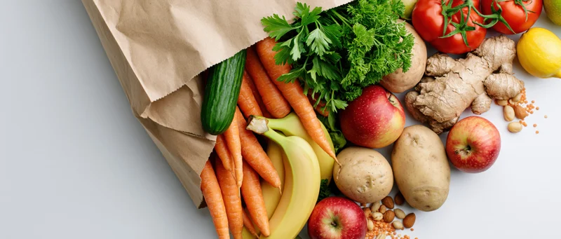 Kraft paper grocery bag tipped over spilling fresh vegetables and fruits on white surface
