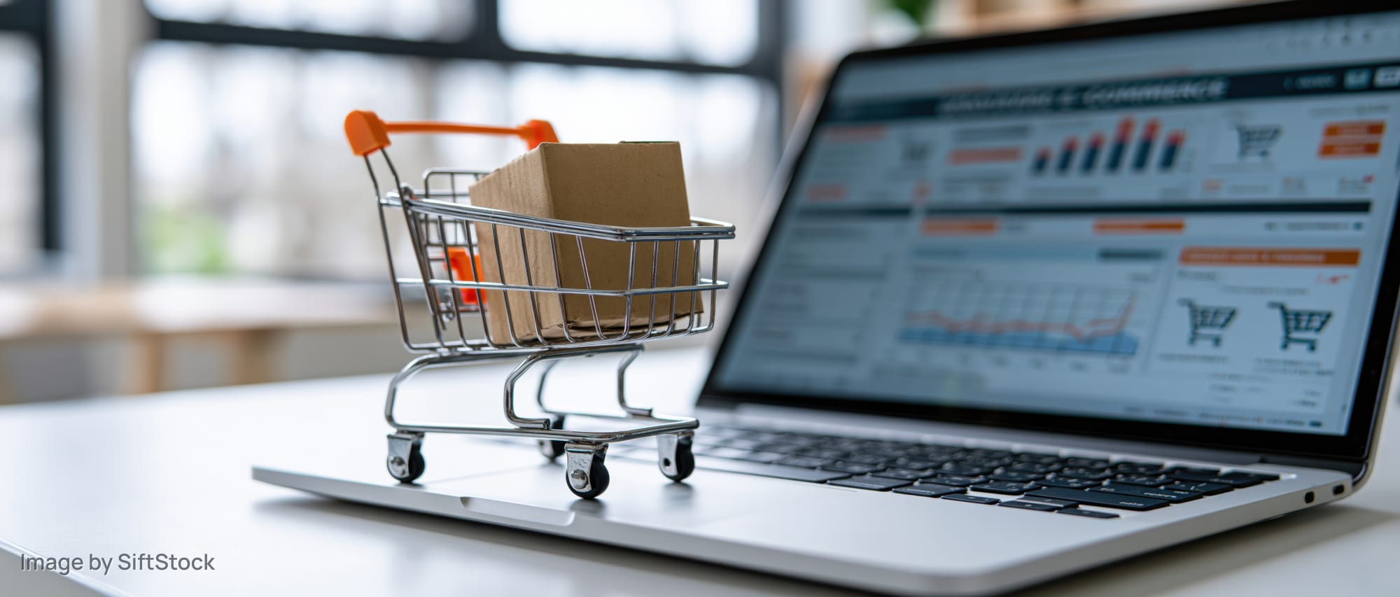 Miniature shopping cart with cardboard box on laptop displaying e-commerce analytics dashboard