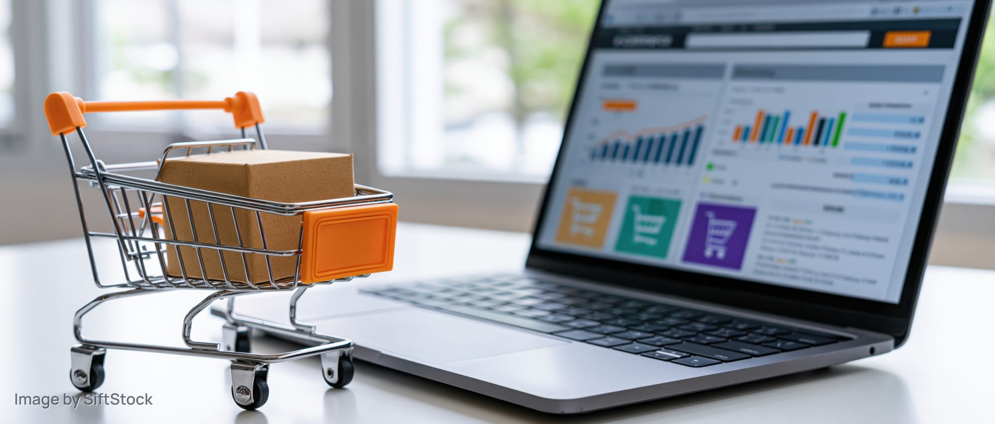 Miniature shopping cart with box beside laptop displaying e-commerce analytics dashboard on desk
