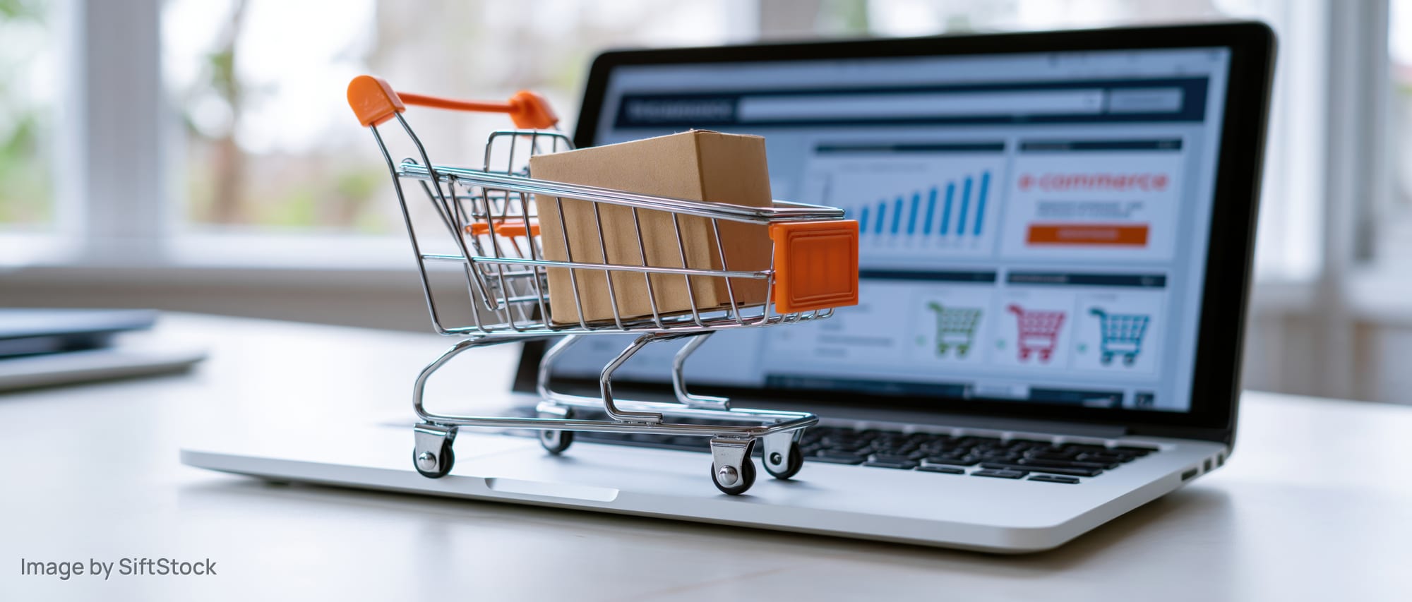 Miniature shopping cart with box on laptop displaying e-commerce dashboard on desk
