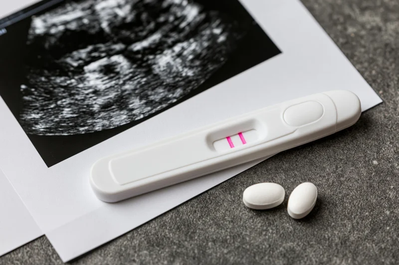 Positive pregnancy test with two pink lines resting on ultrasound scan and white pills on gray surface