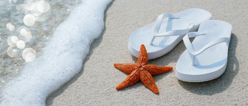 Orange starfish on wet sand beside white flipflops near ocean foam