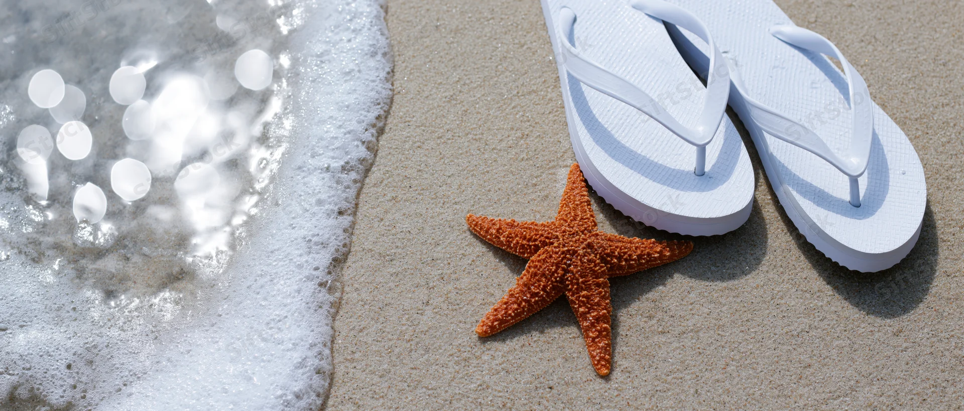 Rust-orange starfish on wet sand beside white flipflops near ocean waves