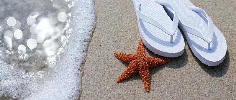 Rust-orange starfish on wet sand beside white flipflops near ocean waves