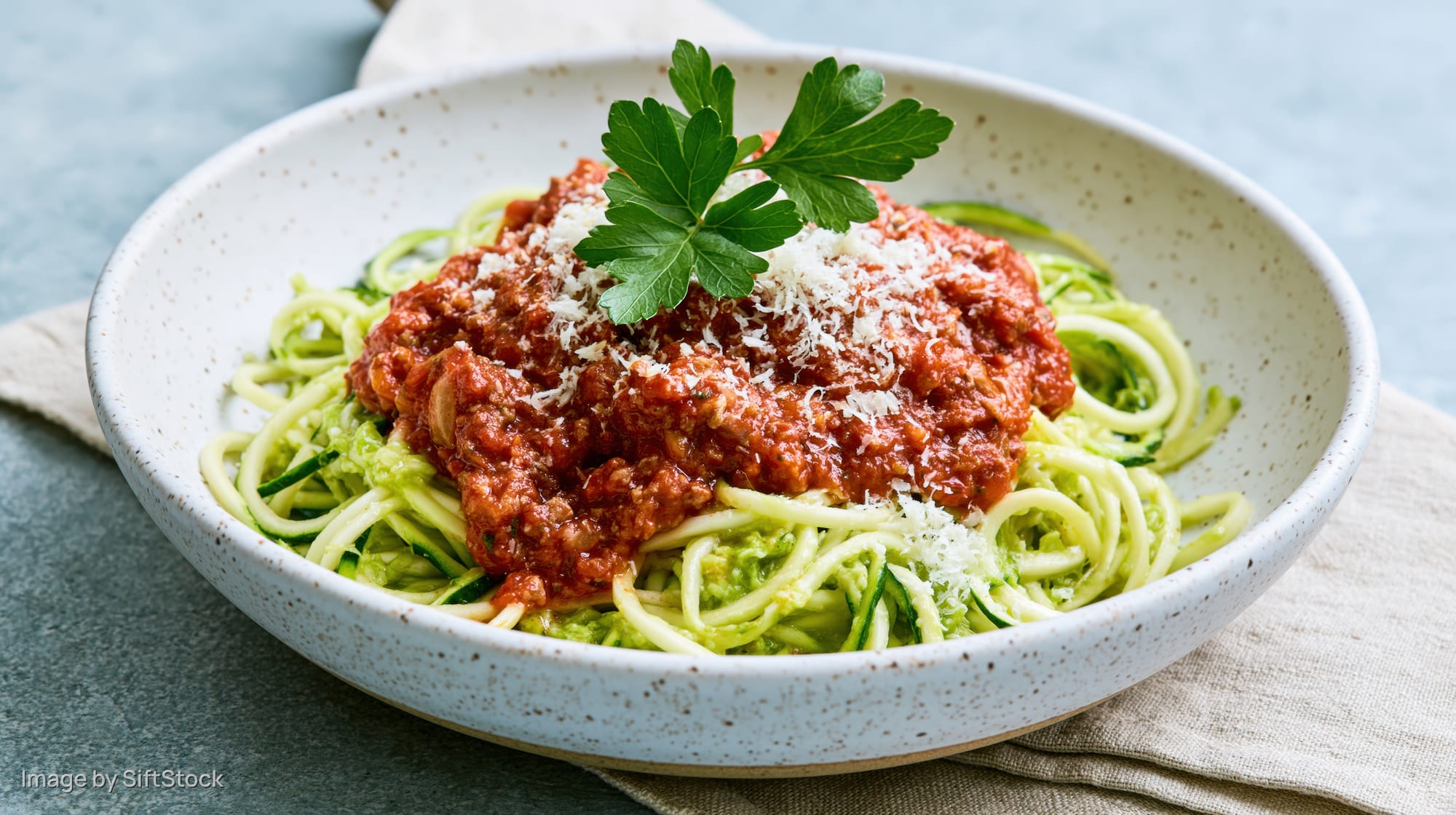 Spiralized zucchini noodles with tomato sauce and parmesan in a white speckled ceramic bowl on a gray surface