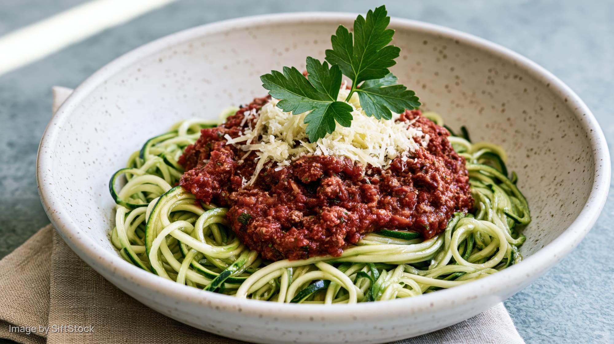 Spiralized zucchini noodles topped with meat sauce and cheese in a white speckled ceramic bowl on a cloth napkin