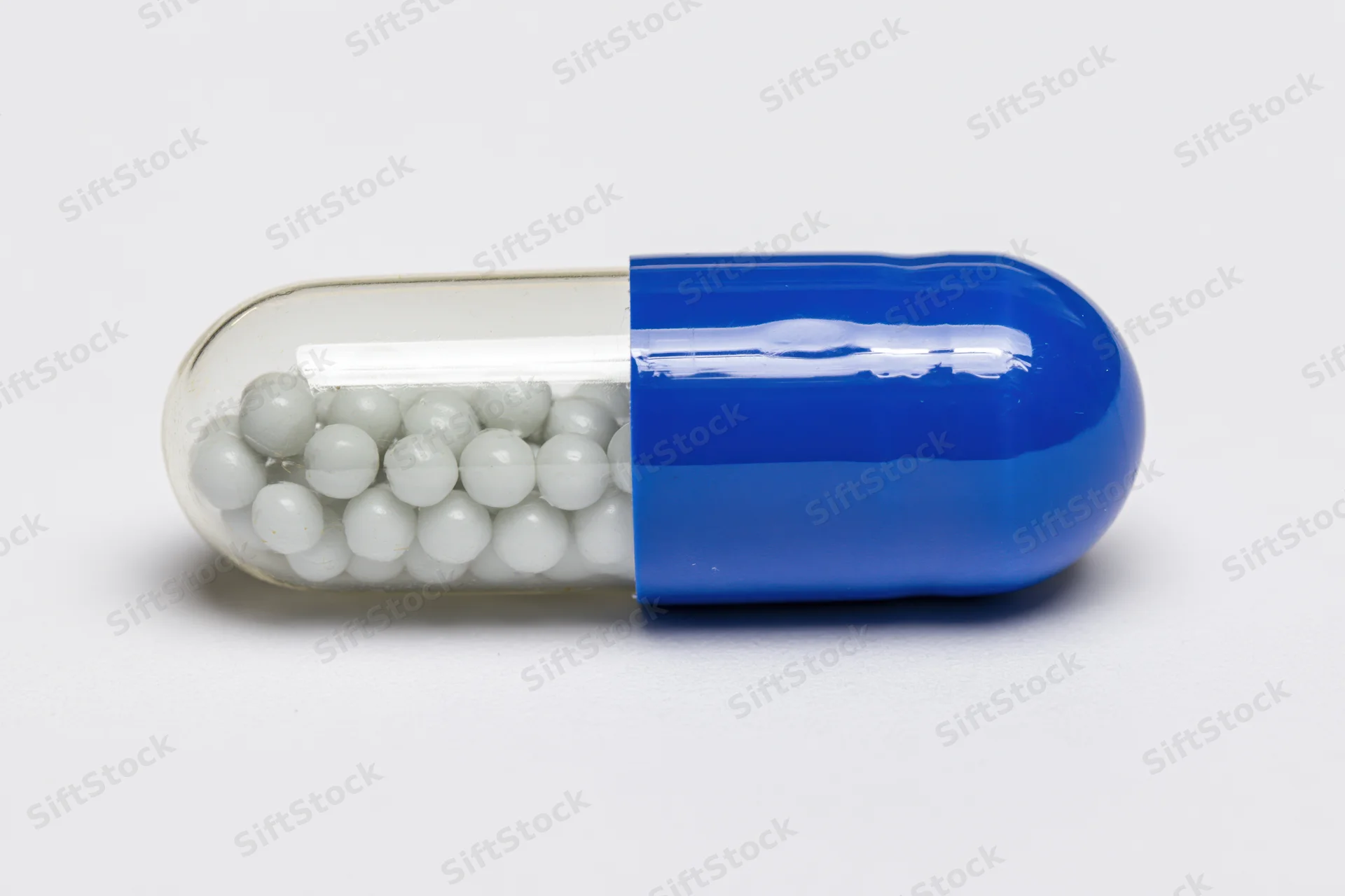 A pharmaceutical capsule with white beads inside, one half blue and one half transparent, lying horizontally on a white surface