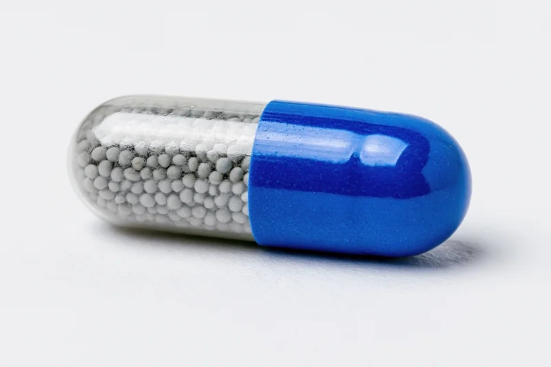 A pharmaceutical capsule with blue and transparent halves containing white beads lying horizontally on a white surface