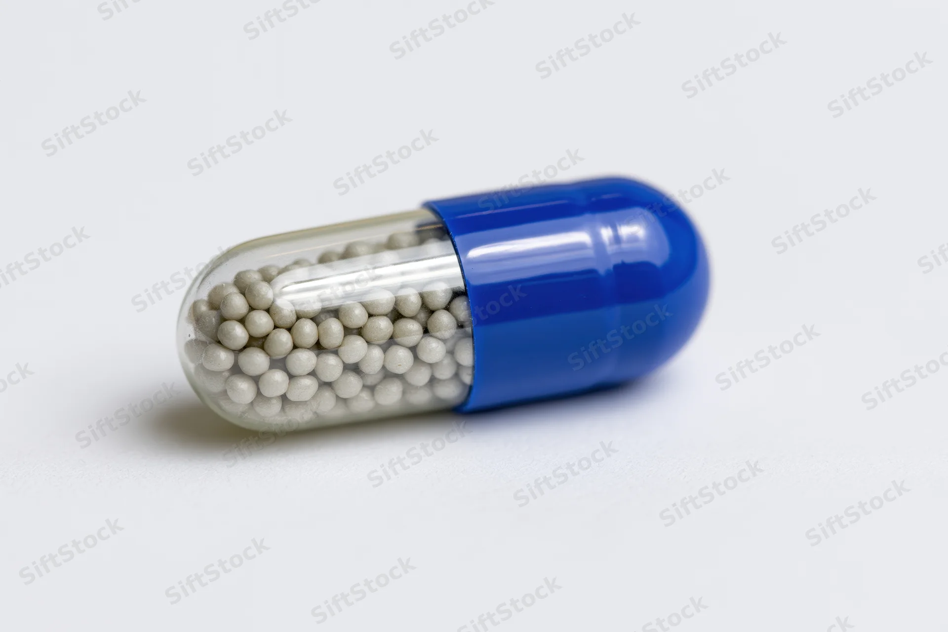 A pharmaceutical capsule with blue and transparent halves filled with white beads lying horizontally on a white surface