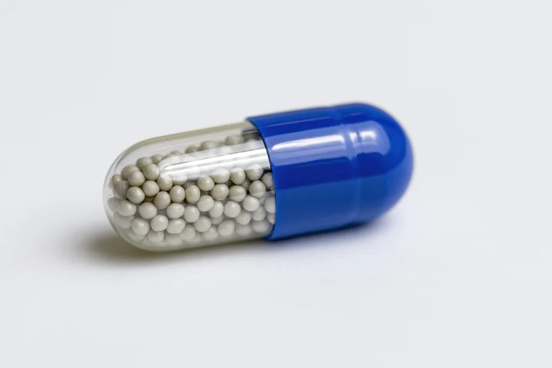 A pharmaceutical capsule with blue and transparent halves filled with white beads lying horizontally on a white surface