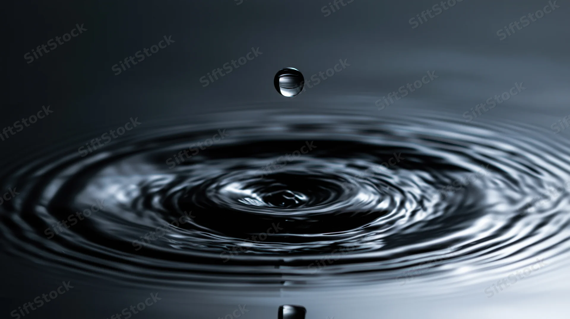 A water droplet suspended above a dark still surface creating concentric ripples
