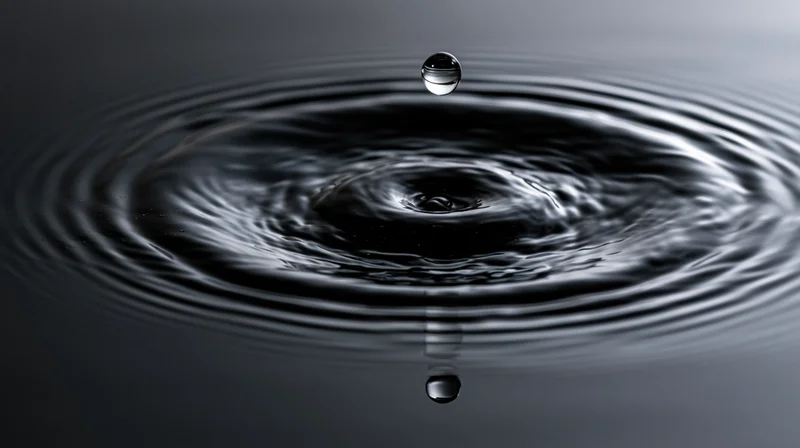 A single water droplet suspended above a dark still water surface creating ripples and reflection