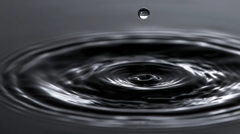 Single water droplet suspended above dark still water surface creating concentric ripples