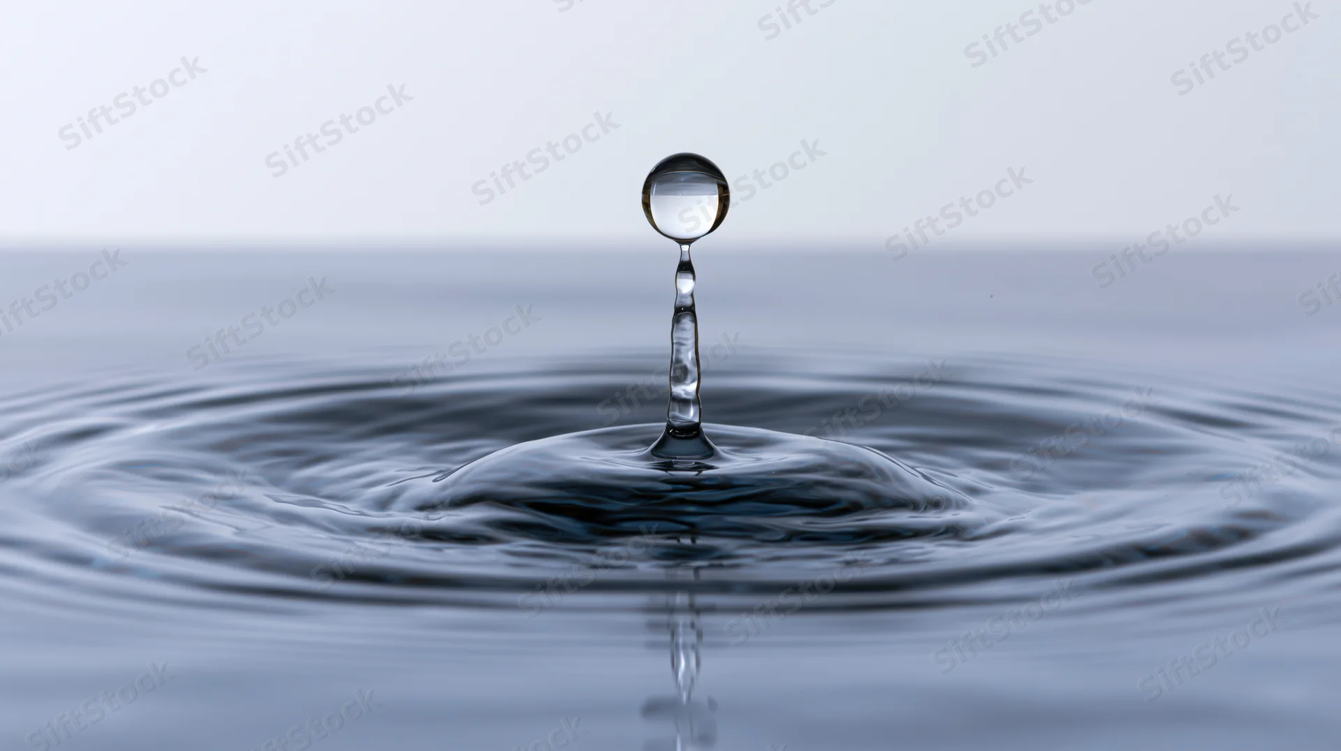 Water droplet suspended above calm surface creating ripples and reflection