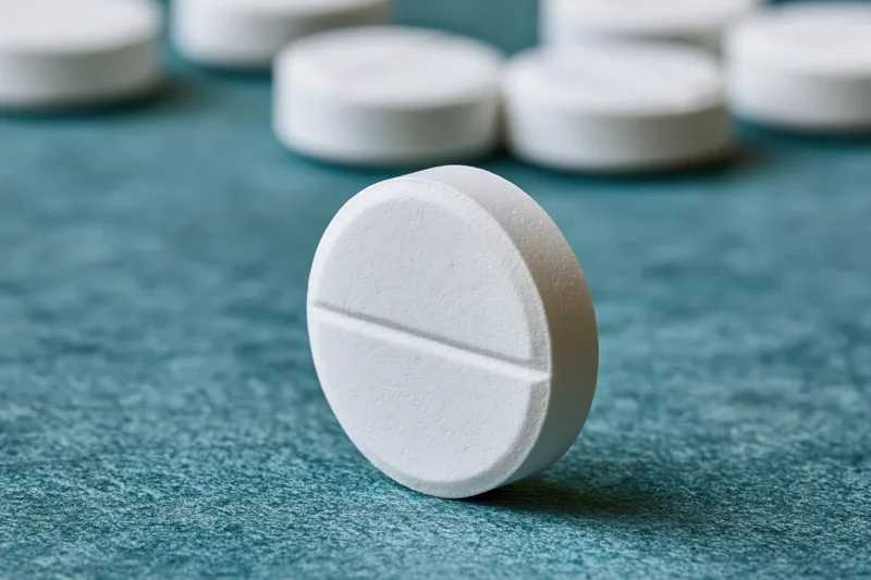Single white scored tablet standing upright on textured teal surface with blurred pills in background
