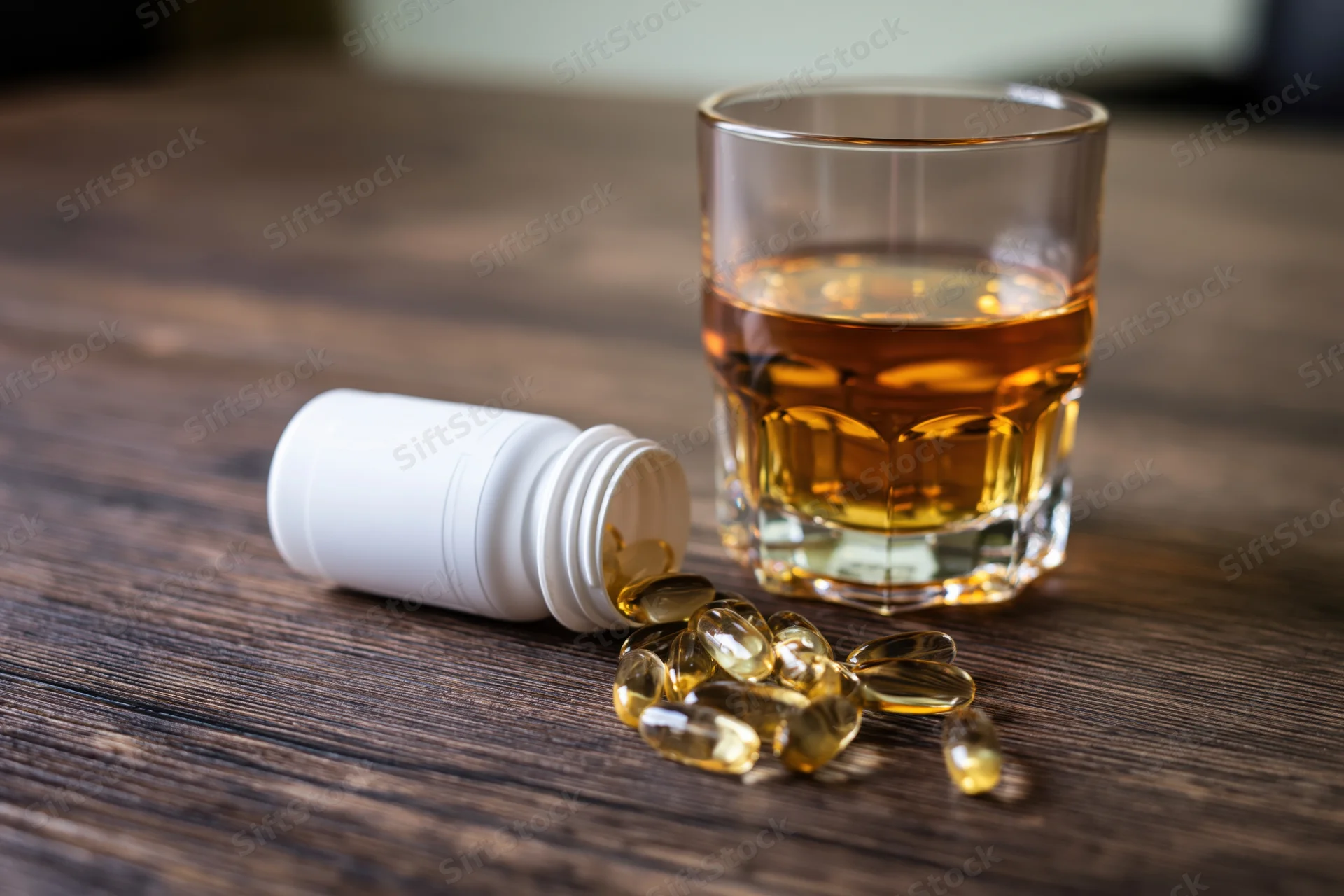 Small glass of amber whiskey beside spilled capsules from white bottle on wooden table