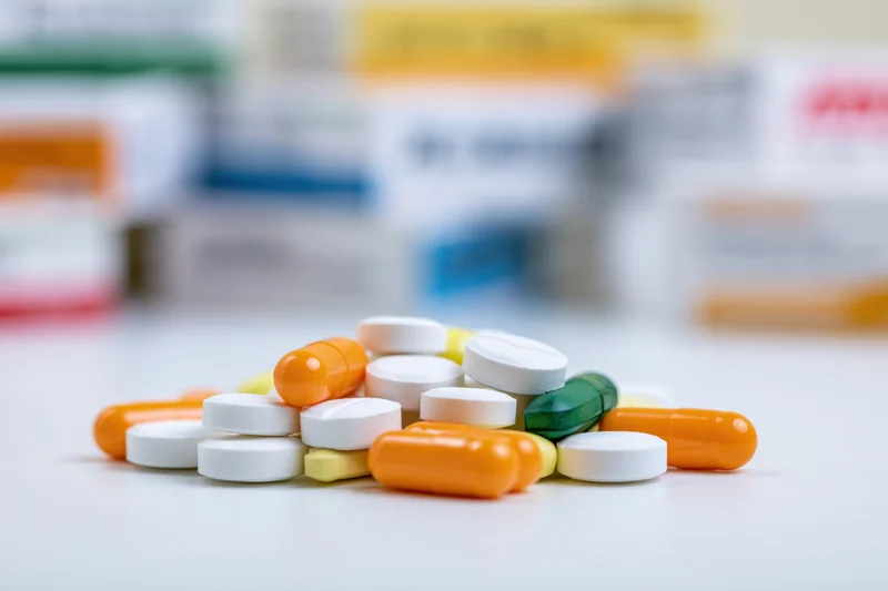 Scattered pile of assorted pills and capsules on a white surface with blurred medication boxes in background
