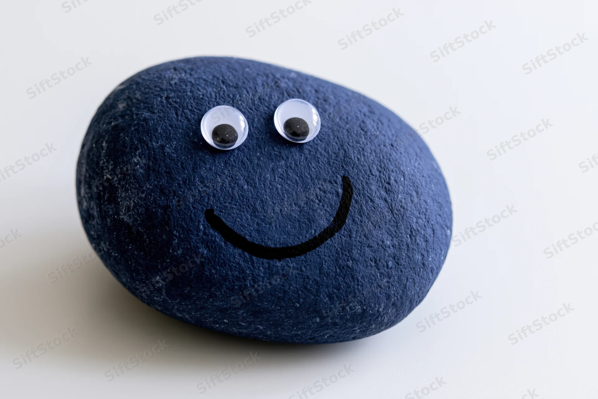 Dark blue painted rock with googly eyes and smiley face on white background