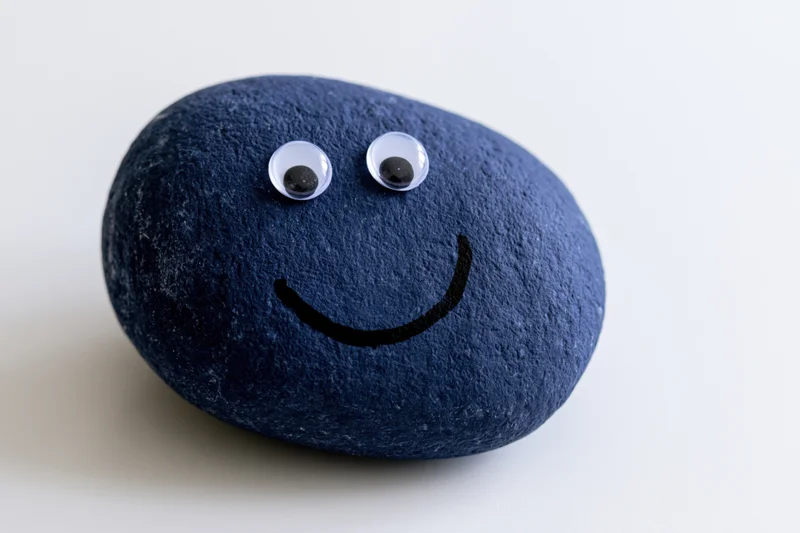 Dark blue painted rock with googly eyes and smiley face on white background