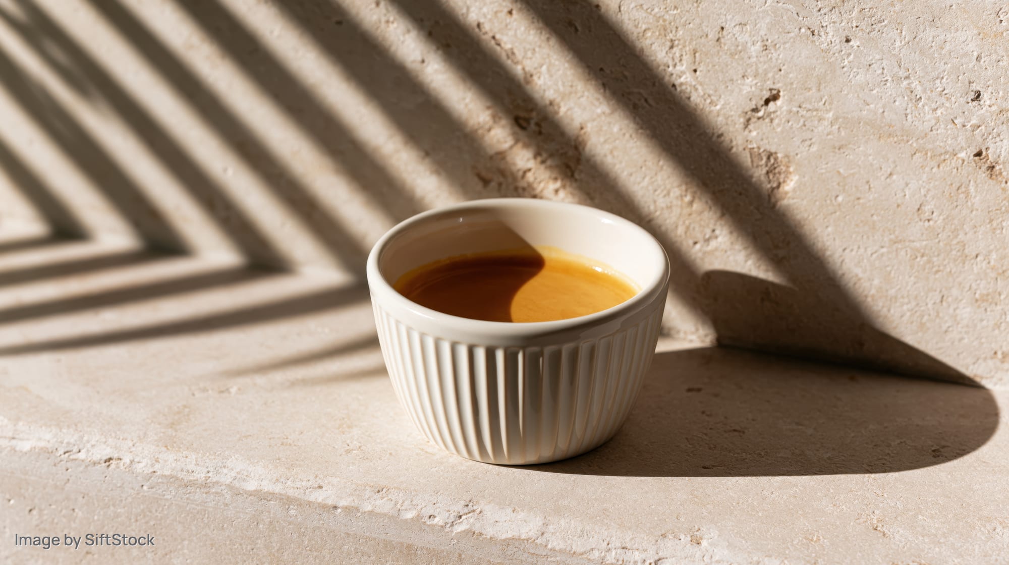 Small white ridged ceramic cup filled with espresso on a textured stone surface with striped sunlight shadows