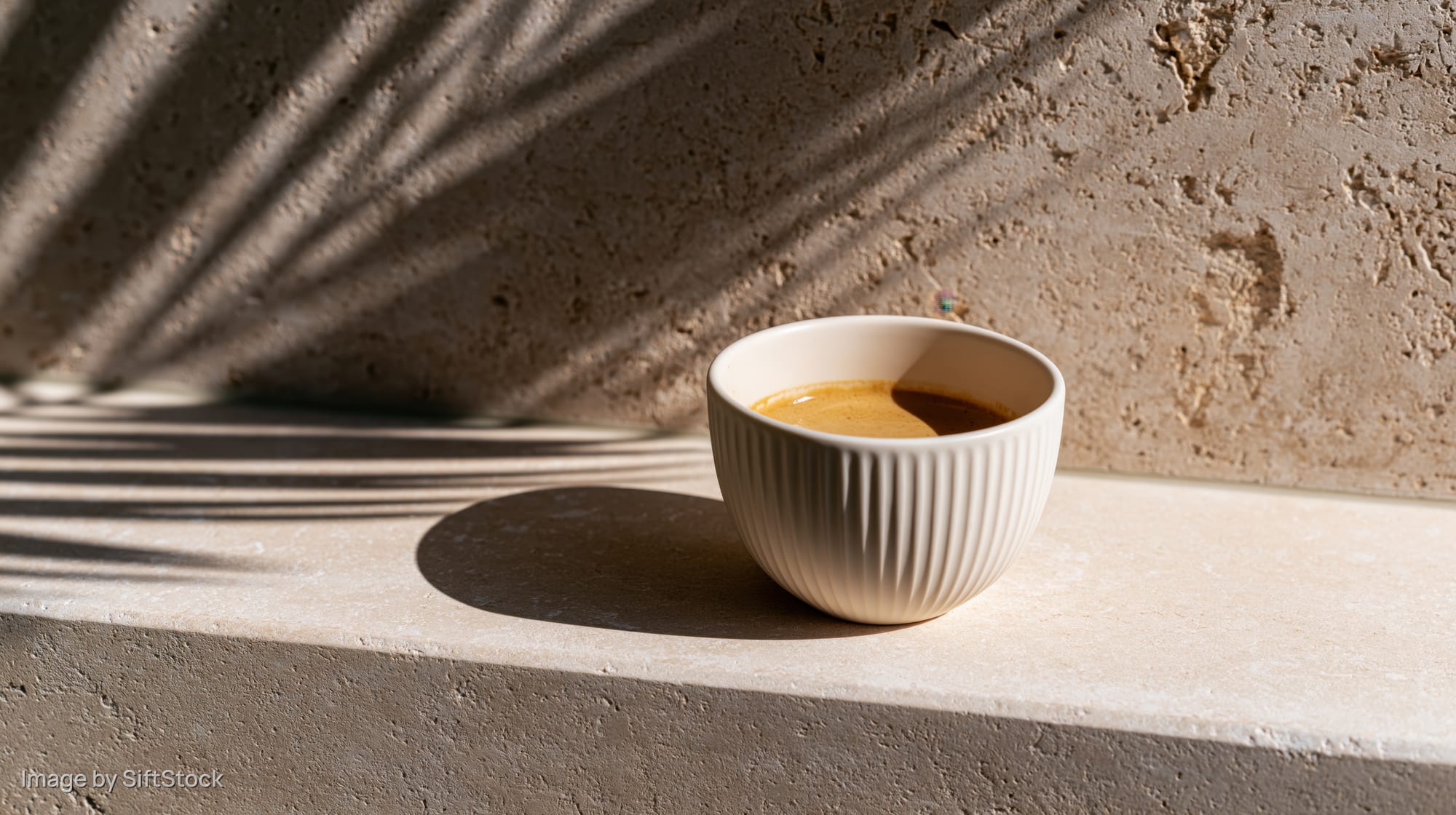 White ridged ceramic cup filled with espresso on a concrete ledge with leaf shadows in natural sunlight