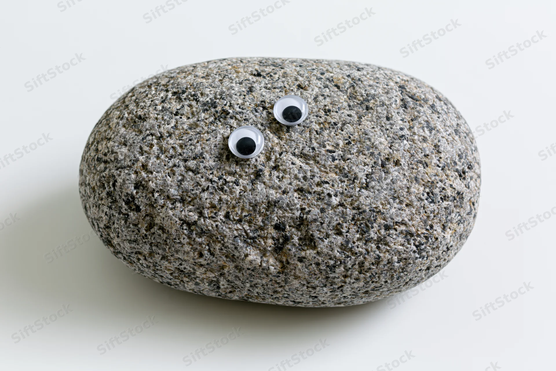 Smooth oval grey river rock with two small googly craft eyes on white background