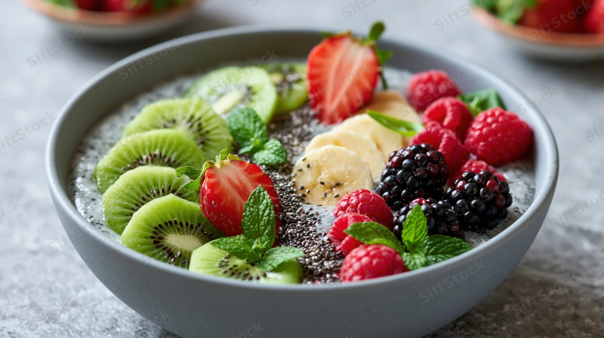 Smoothie bowl topped with sliced kiwi, strawberries, banana, raspberries, blackberries, chia seeds, and mint leaves on a grey ceramic bowl on a stone countertop