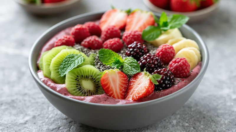 Smoothie bowl topped with sliced fruits and mint in a matte grey ceramic bowl on a stone surface