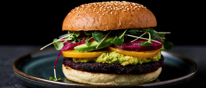Stacked vegan burger with black bean patty, avocado, beet, and microgreens on a seeded brioche bun, served on a dark plate
