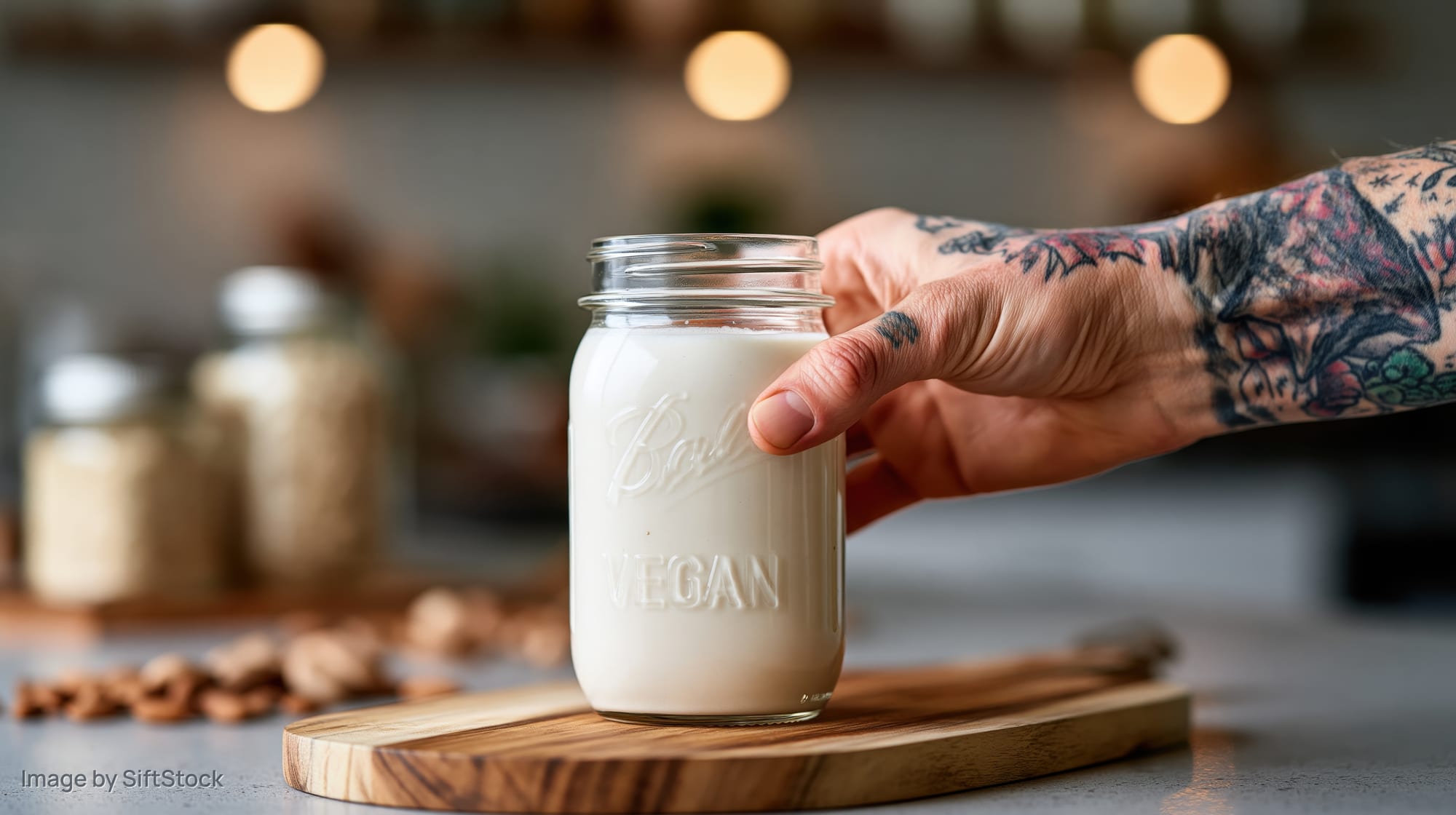 Tattooed hand placing a glass mason jar filled with creamy vegan milk on a wooden cutting board in a kitchen