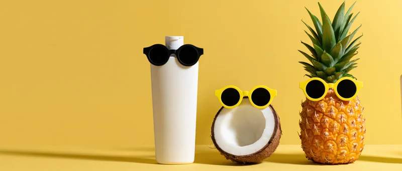 Pineapple, coconut, and sunscreen bottle wearing sunglasses arranged on yellow surface