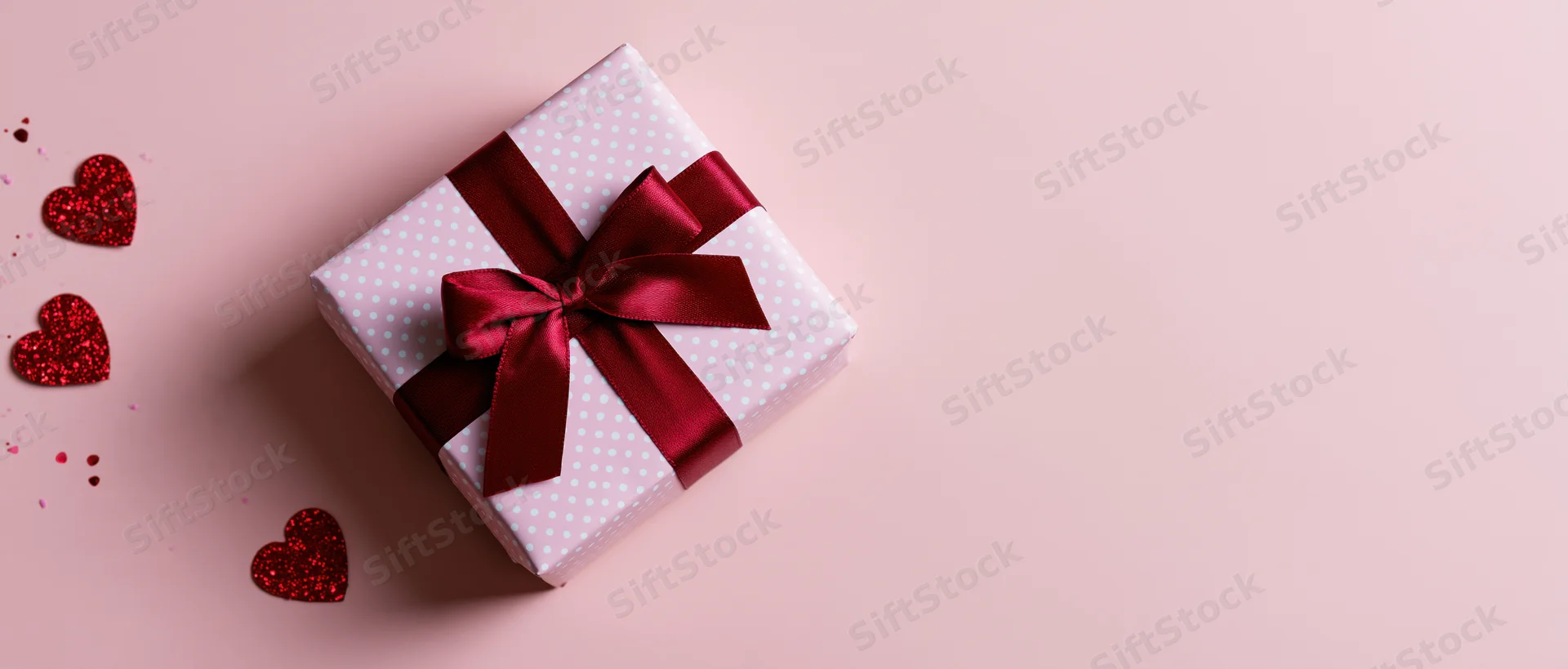 Wrapped gift box with red ribbon and glitter hearts on pink background