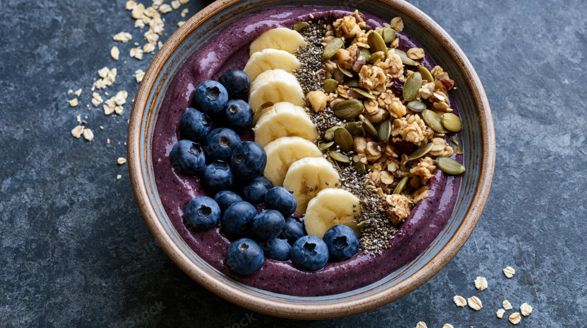 Acai smoothie bowl topped with blueberries, banana slices, granola, and seeds on a dark textured surface