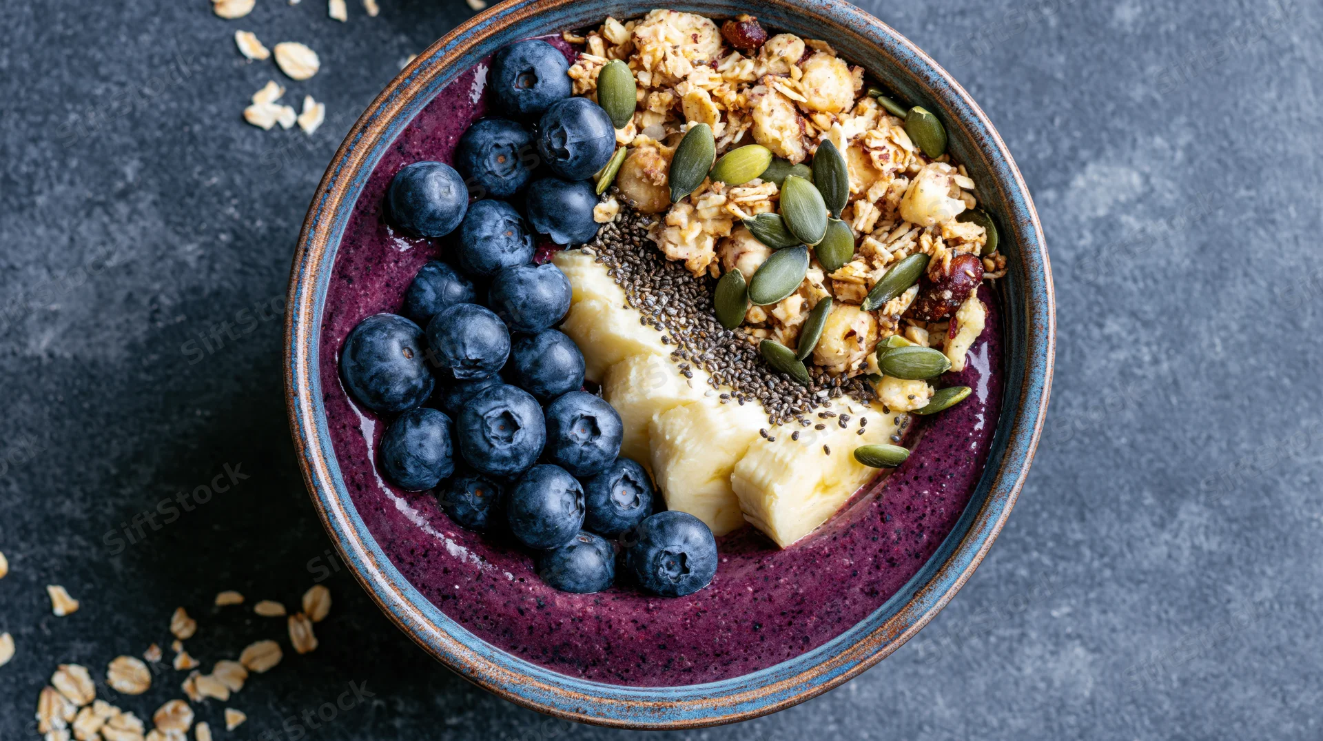 Acai smoothie bowl topped with blueberries, banana, granola, and seeds on dark stone surface
