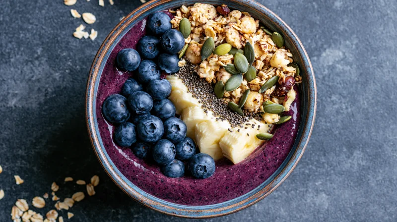 Acai smoothie bowl topped with blueberries, banana, granola, and seeds on dark stone surface