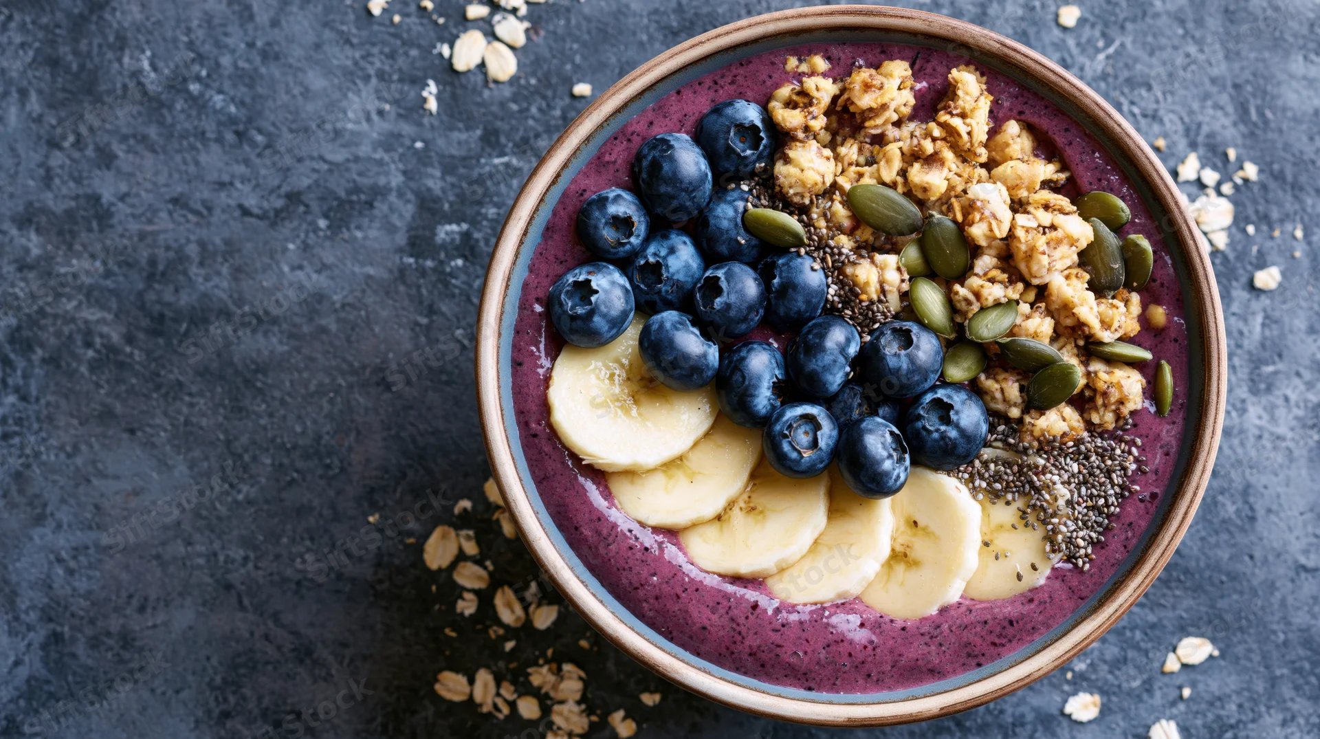 Acai smoothie bowl topped with blueberries, banana slices, granola, and seeds on dark textured surface